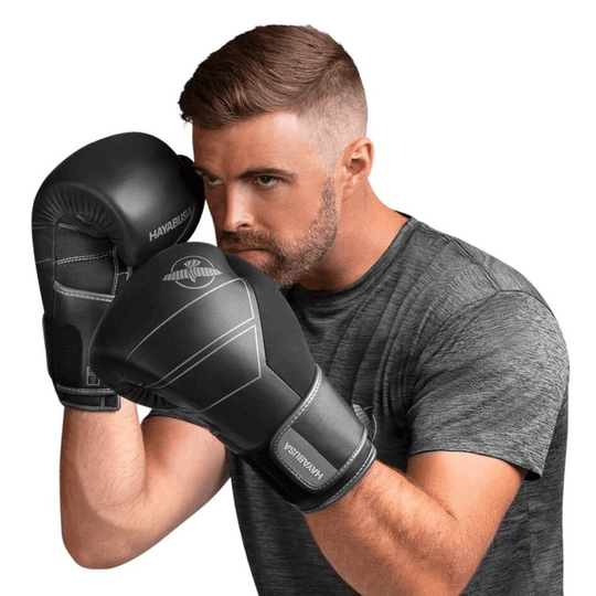 Een man met een donkergrijs shirt poseert met de Hayabusa Bokshandschoenen S4 Leather in zwart, klaar in een bokshouding met de handschoenen ter hoogte van zijn gezicht.