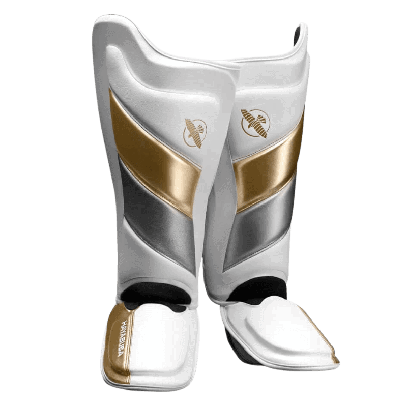 Een paar Hayabusa T3 Striking Scheenbeschermers in wit en goud, van voren bekeken. Ze hebben een strak ontwerp met het gouden Hayabusa valk-logo bovenop.