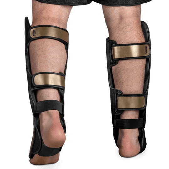 Een zijaanzicht van een persoon die Hayabusa T3 Striking Scheenbeschermers in zwart en goud draagt, waarbij de structuur en de verstelbare gouden banden aan de achterzijde zichtbaar zijn.