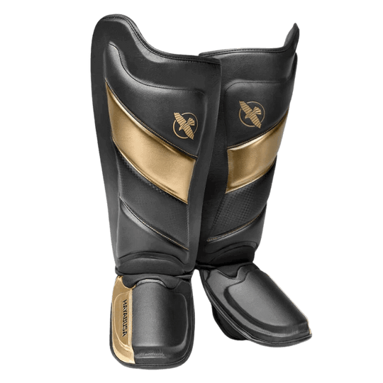 Een paar Hayabusa T3 Striking Scheenbeschermers in zwart en goud, frontaal beeld, met het gouden Hayabusa valk-logo op het bovenste deel.