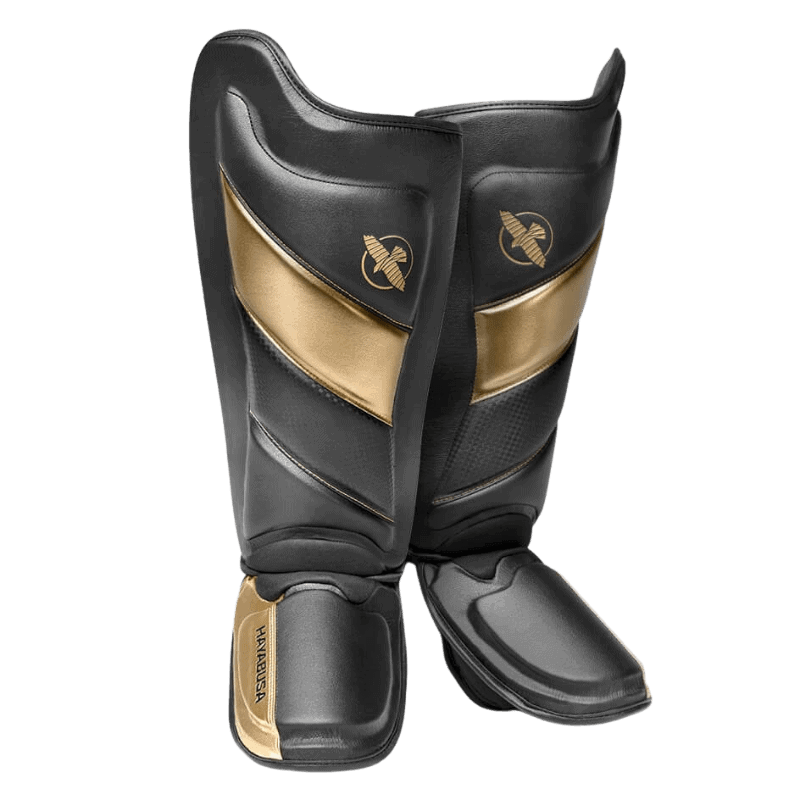 Een paar Hayabusa T3 Striking Scheenbeschermers in zwart en goud, frontaal beeld, met het gouden Hayabusa valk-logo op het bovenste deel.