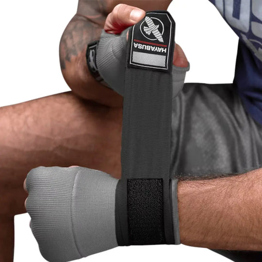 Close-up van een man met getatoeëerde arm die een grijze Hayabusa binnenhandschoen draagt en de lange, grijze polsbandage met zwarte klittenband strak om zijn pols wikkelt.