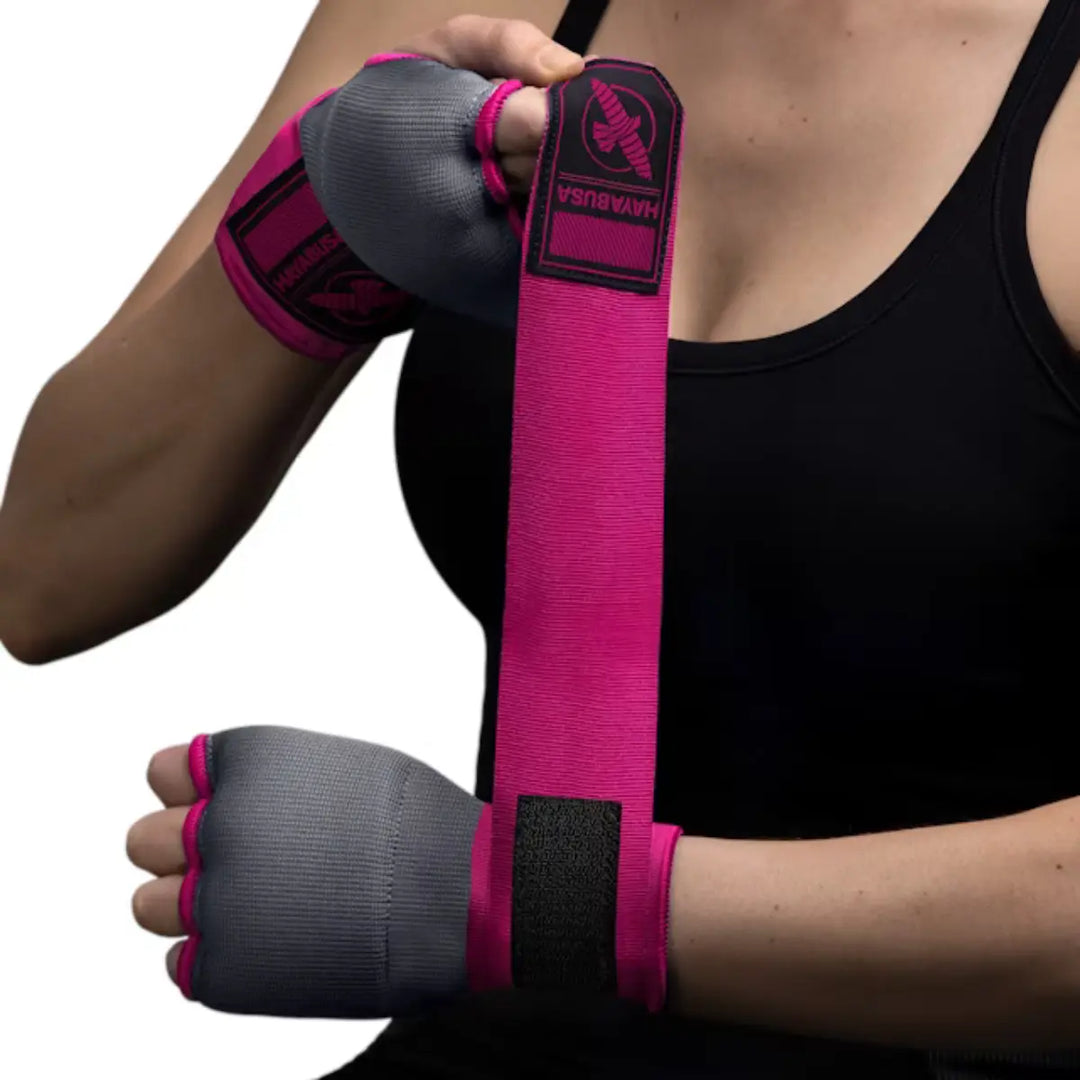 Een vrouw in een zwarte sporttop wikkelt haar handen in met felroze Hayabusa binnenhandschoenen/handwraps (quick wraps) met grijze vingerloze handschoen en een zwart logo.