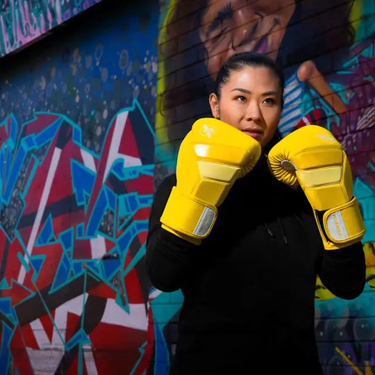 Vechter met gele Hayabusa T3 bokshandschoenen poseert in gevechtshouding voor een muur met kleurrijke graffiti.