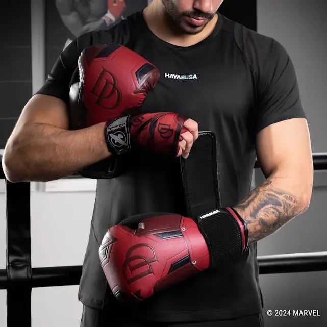 Model in een boksring dat de rode Hayabusa Daredevil bokshandschoenen aandoet, waarbij de polsband met klittenband en het grote DD-logo zichtbaar zijn.