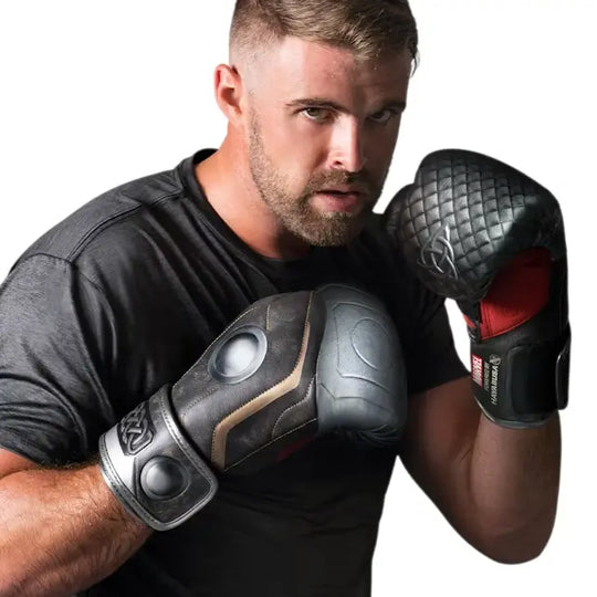 Een man met een baard en een zwart T-shirt poseert in een vechthouding terwijl hij bokshandschoenen draagt die geïnspireerd zijn op het pantser van Marvel's Thor. De handschoenen zijn zwart en grijs en hebben gedetailleerde, gegraveerde ontwerpen op de manchetten.