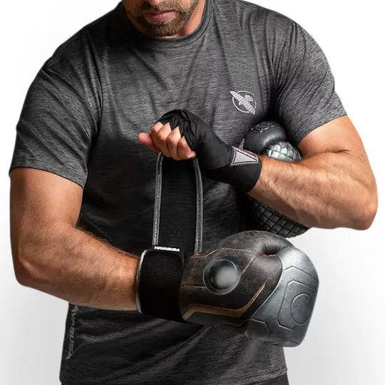 Een atleet in een grijs sportshirt houdt een paar zwarte Thor-geïnspireerde bokshandschoenen vast. Zijn handen zijn ingezwachteld met zwarte handwraps en de handschoenen worden vastgehouden via hun Dual-X sluiting.