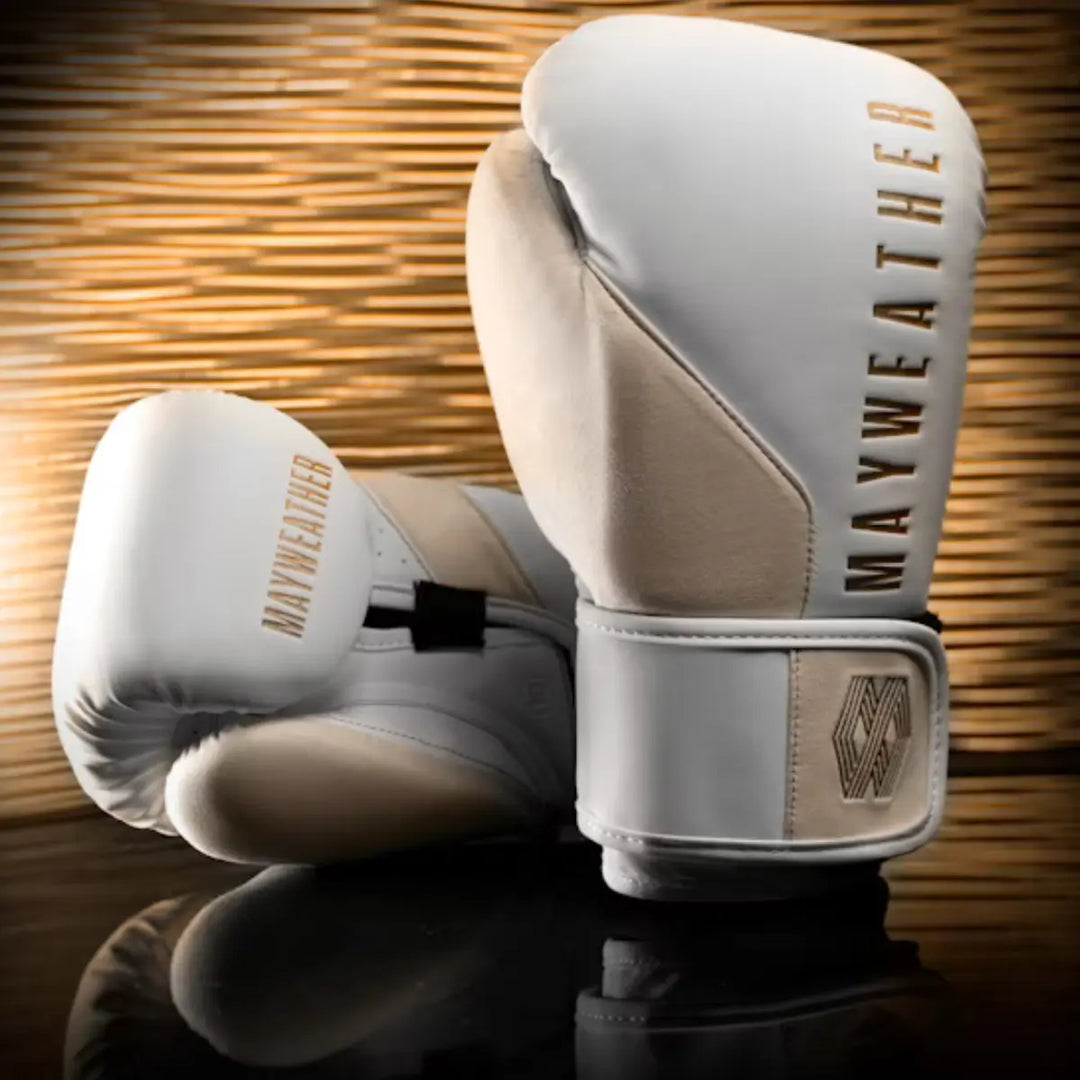 Een paar witte en beige boks- of MMA-handschoenen met het goudkleurige Mayweather-logo staat tegen een reflecterende donkere achtergrond met gouden lichteffecten. De handschoenen hebben een strak ontwerp en een geometrisch 'M'-logo op de pols.