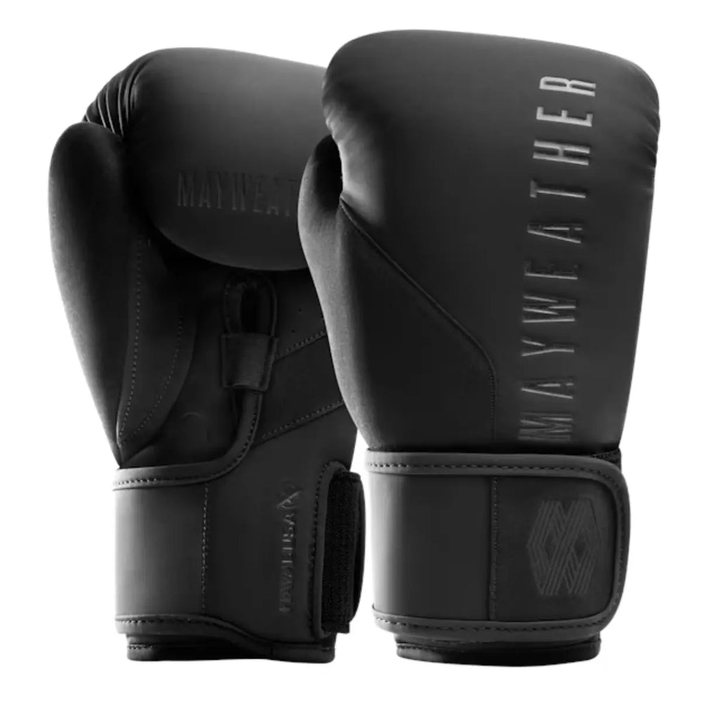 Een paar matzwarte boks- of MMA-handschoenen met het Mayweather logo in een donkere, glanzende afwerking staat rechtop. De handschoenen hebben een zwart-op-zwart 'M'-logo op de polsband en het Hayabusa logo is subtiel zichtbaar op de linkerhandschoen.