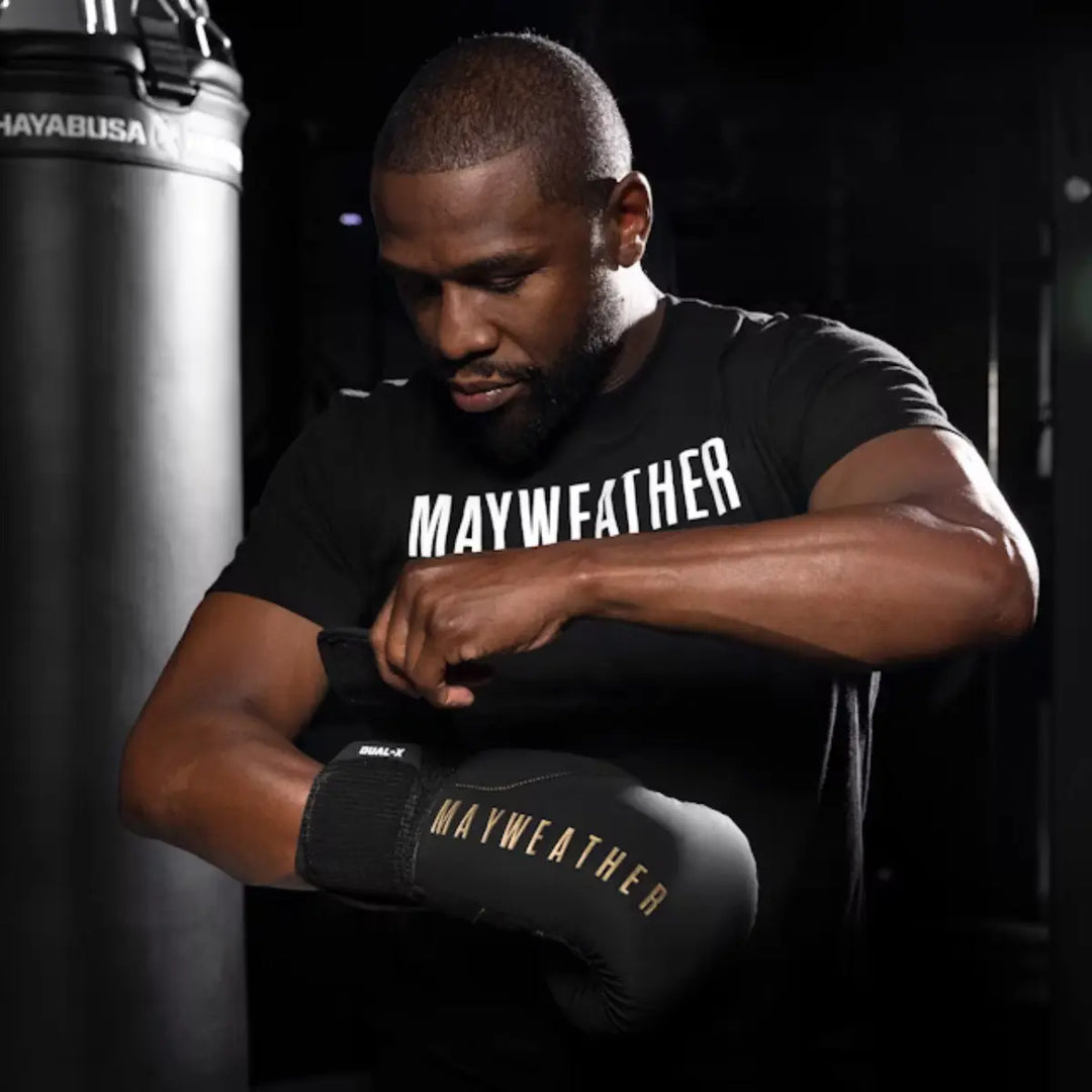 Bokser in zwarte Mayweather-handschoenen maakt zich klaar voor training in een donkere gym, met focus en concentratie zichtbaar in zijn houding.