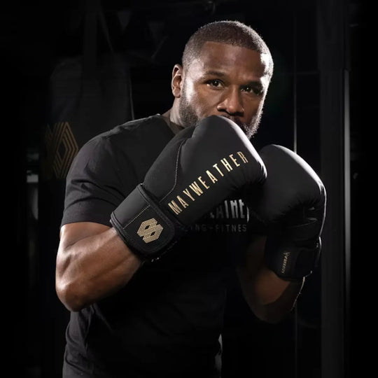 Bokser in gevechtshouding met zwarte Mayweather bokshandschoenen met gouden details, klaar voor actie in een donkere trainingsruimte.