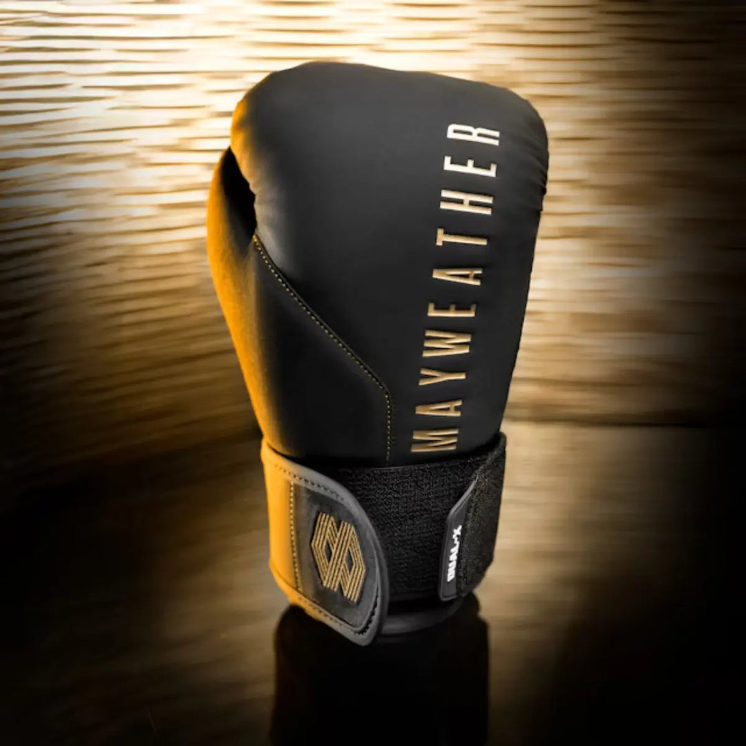Zwarte Mayweather bokshandschoen met gouden opdruk en luxe afwerking, glanzend belicht tegen een gouden achtergrond voor een exclusieve uitstraling.