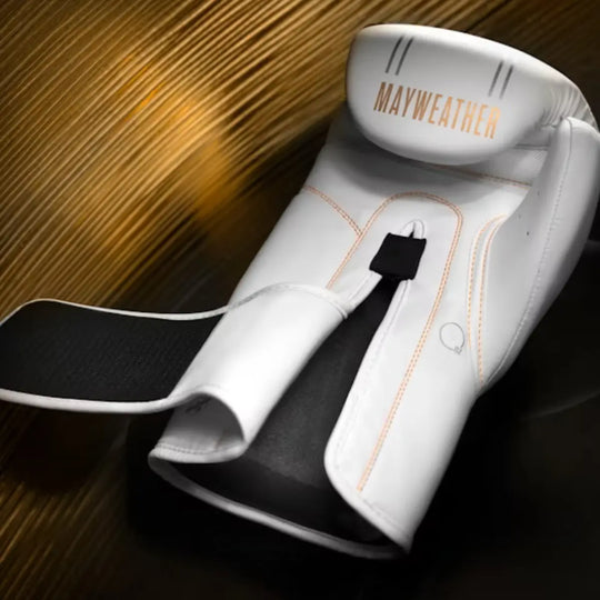 Een enkele witte boks- of trainingshandschoen met het Mayweather logo in goudkleur op de manchet ligt op een donkere ondergrond. De handschoen heeft oranje stiksels en een zwarte klittenbandsluiting.