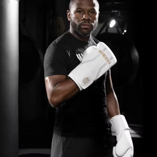 Bokser Floyd Mayweather kijkt direct in de camera, terwijl hij een zwart T-shirt en witte boks- of trainingshandschoenen draagt. De handschoenen, met het gouden Mayweather-logo en zilveren details, steken af tegen de donkere fitnessruimte op de achtergrond.