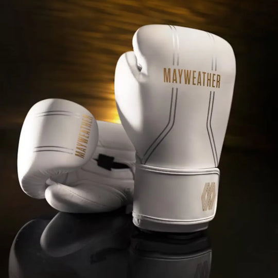 Een paar witte boks- of MMA-handschoenen met het goudkleurige Mayweather-logo staat prominent op een reflecterende donkere en gouden achtergrond. De handschoenen hebben zilveren streepdetails en een gouden geometrisch logo op de polsband.