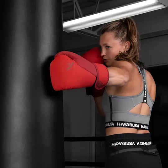 Vrouwelijke kickbokser met rode Hayabusa T3 bokshandschoenen traint op een bokszak in de sportschool, gefocust op haar stoottechniek.