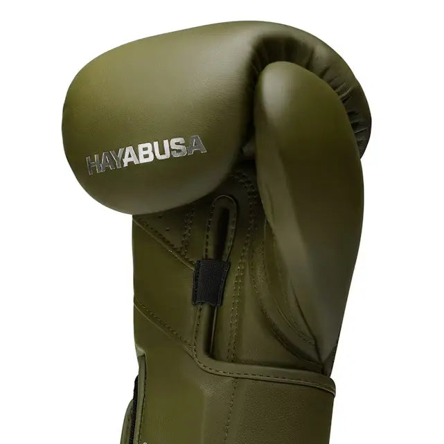 Een close-up toont de zijkant van een olijfgroene Hayabusa bokshandschoen met het zilveren Hayabusa logo prominent op de knokkel. De handschoen heeft een slanke vormgeving en een duidelijke stiklijn langs de duim.