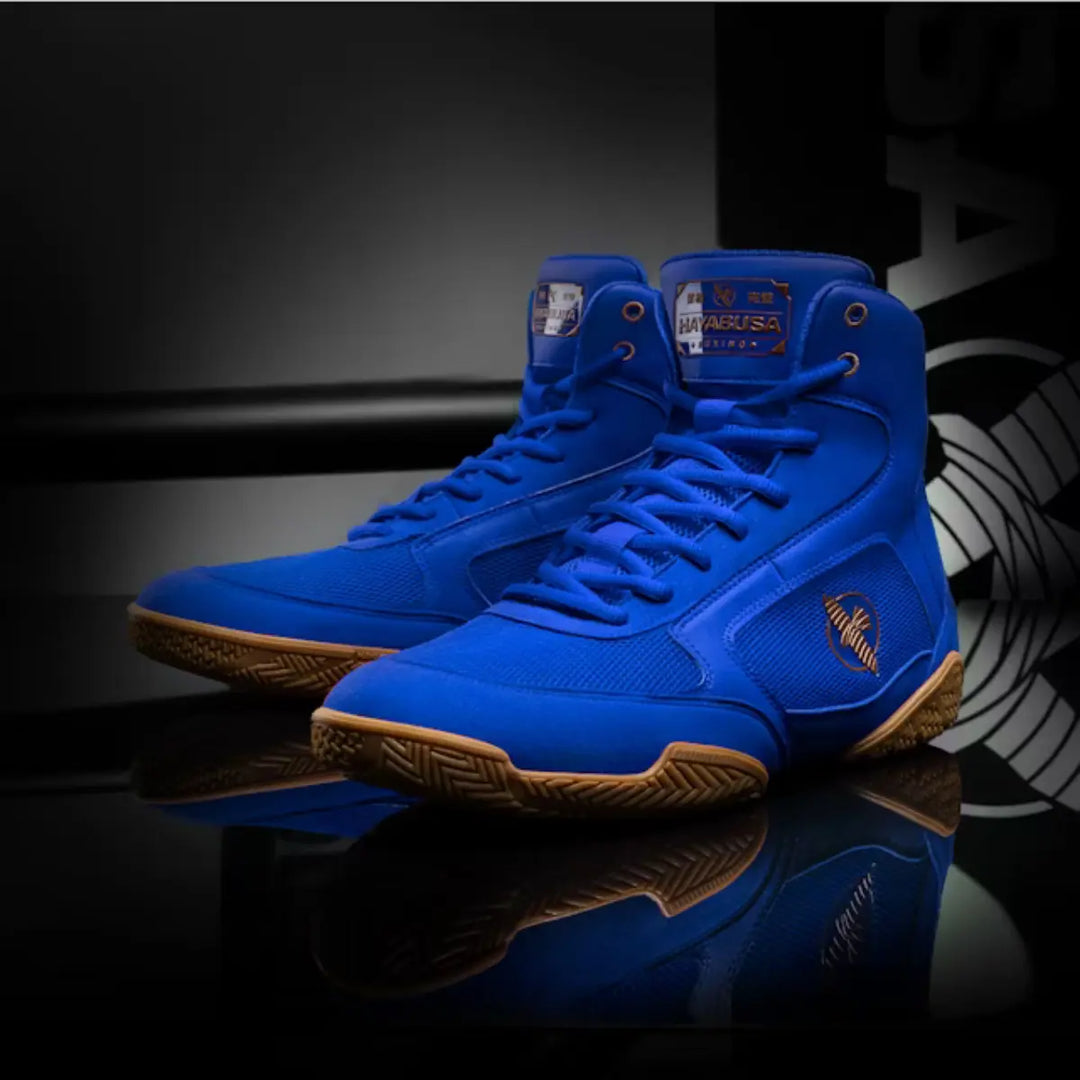Blauwe Hayabusa boksschoenen met bruin/oranje zolen en details, gepresenteerd op een glanzende zwarte ondergrond.
