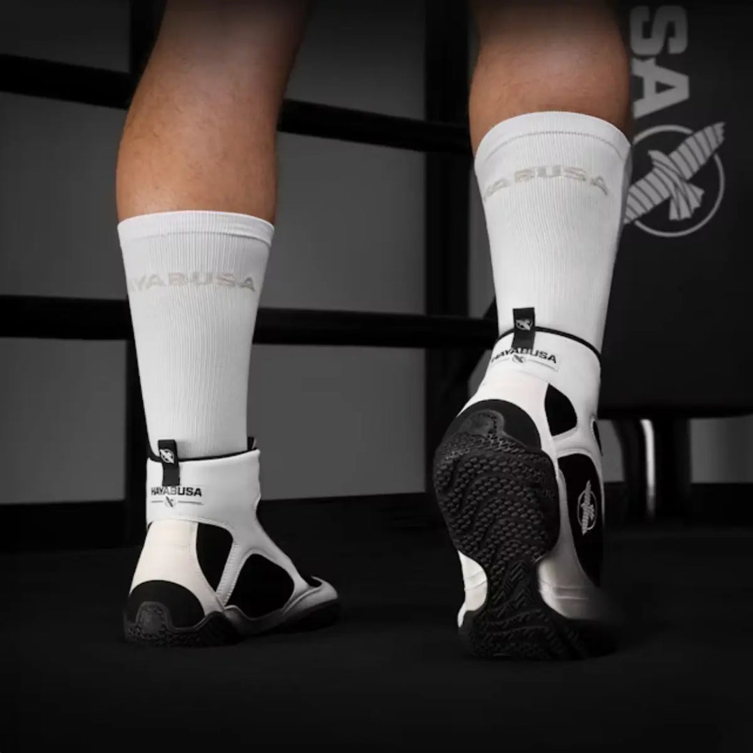 Achteraanzicht van witte en zwarte Hayabusa boksschoenen gedragen met witte sokken in gymomgeving met Hayabusa logo.