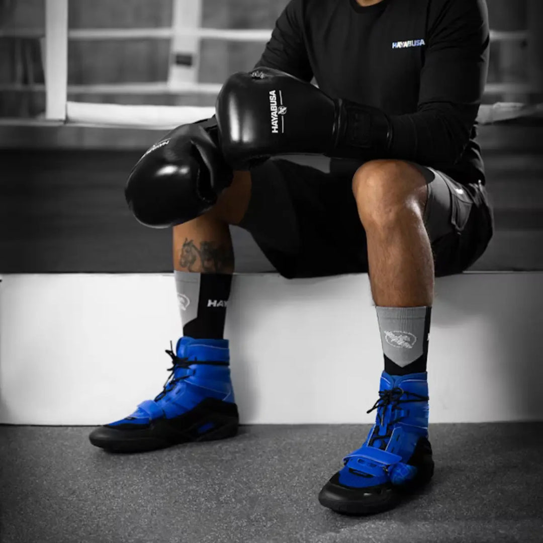 Bokser zittend in de gym met zwarte Hayabusa bokshandschoenen en blauwe Hayabusa boksschoenen, ontworpen voor stabiliteit, wendbaarheid en ondersteuning tijdens intensieve bokstrainingen.
