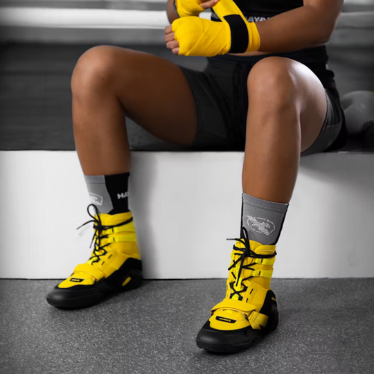 Een bokser zit op de rand van de boksruimte met felgele Hayabusa boksschoenen en bijpassende handwraps. De schoenen bieden stabiliteit, grip en ondersteuning tijdens intensieve bokstrainingen.