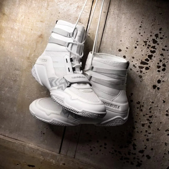 Een paar witte Hayabusa boksschoenen hangt aan veters tegen een ruwe muur. De schoenen hebben een moderne, hoge snit met stevige zool en ademend bovenwerk voor optimale grip en ondersteuning tijdens bokstraining.