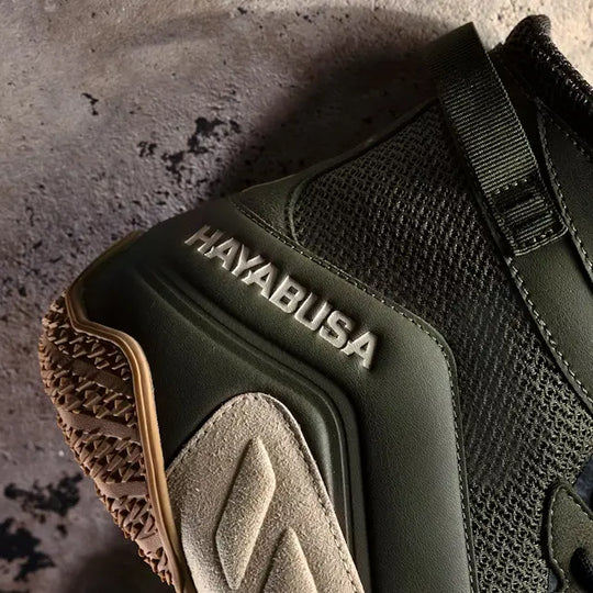 Close-up van het hielgedeelte van een Hayabusa boksschoen in olijfgroen met mesh, suede en rubberen zool, geplaatst op een ruwe betonnen ondergrond.