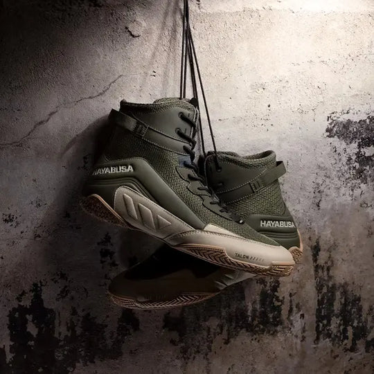 Olijfgroene Hayabusa boksschoenen met beige accenten, hangend aan de veters tegen een verweerde, ruwe betonnen muur.