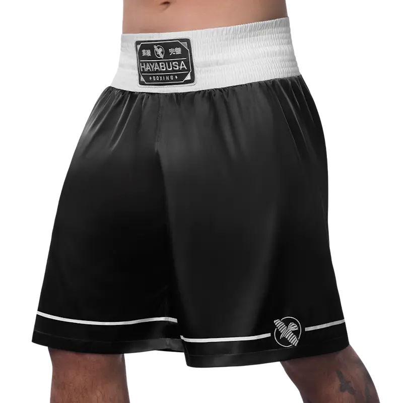 Front left view of black Hayabusa boxing shorts featuring a wide white elastic waistband with stitched Hayabusa Boxing patch. The smooth, lightweight fabric, white piping along the hem, and subtle Hayabusa logo on the leg highlight freedom of movement, comfort, and a clean, professional boxing look.
