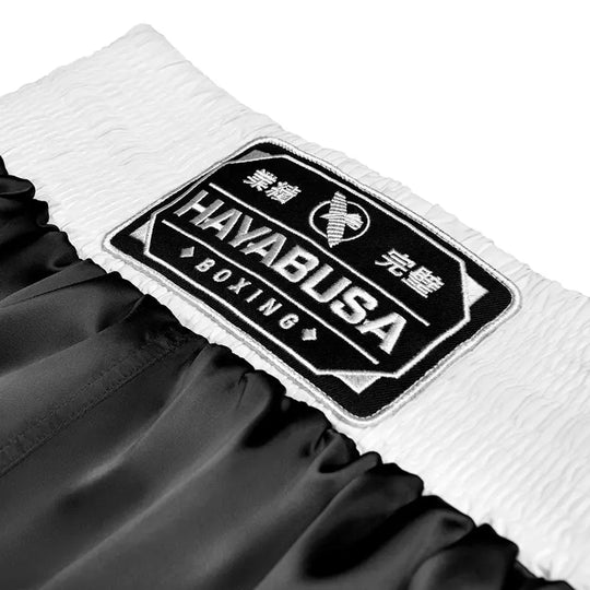 Close-up of the waistband patch on black Hayabusa boxing shorts, featuring the embroidered Hayabusa Boxing logo on a wide white elastic waistband. The detailed stitching and contrast between the black satin fabric and white waistband highlight a premium finish and a secure, traditional boxing fit.