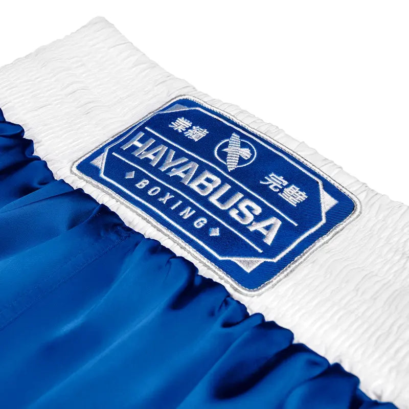 Close-up of the waistband patch on blue Hayabusa boxing shorts, featuring the embroidered Hayabusa Boxing logo on a wide white elastic band. The detailed stitching and premium finish highlight durability and a secure fit around the waist. This waistband construction offers comfort and stability, designed for boxers during intensive training and sparring sessions.