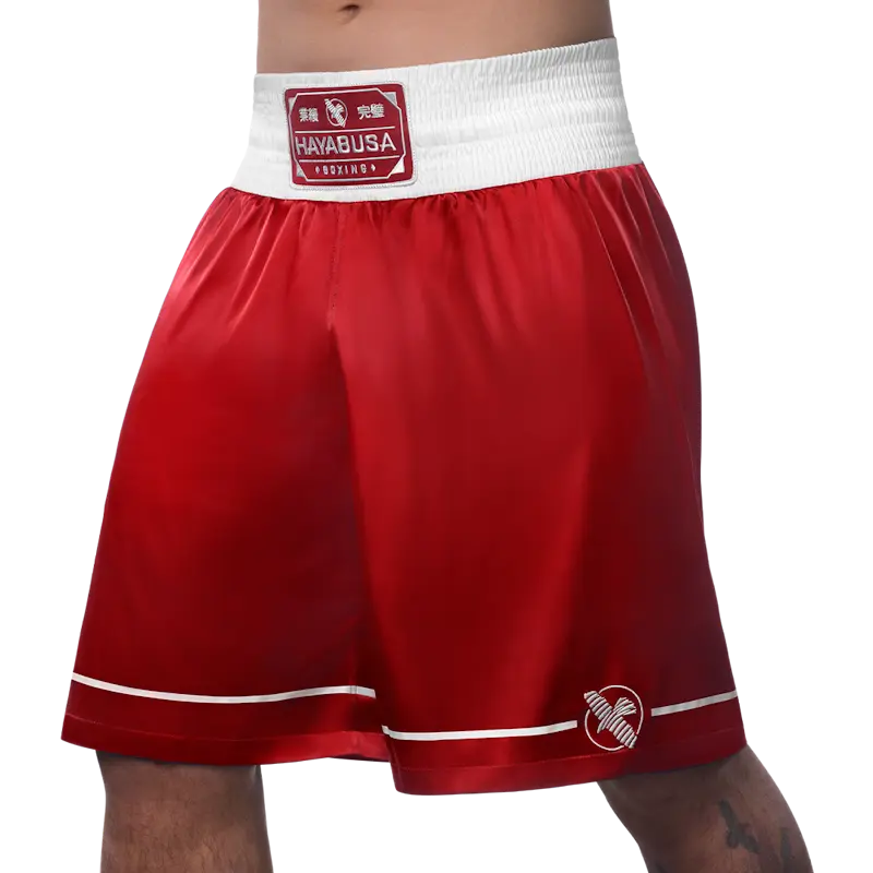 Front left view of red Hayabusa boxing shorts with a wide white elastic waistband and red Hayabusa Boxing patch at the center. The smooth satin fabric features thin white piping along the hem and an embroidered Hayabusa logo on the lower leg. The loose fit emphasizes comfort, mobility, and a classic boxing look for training and gym use.