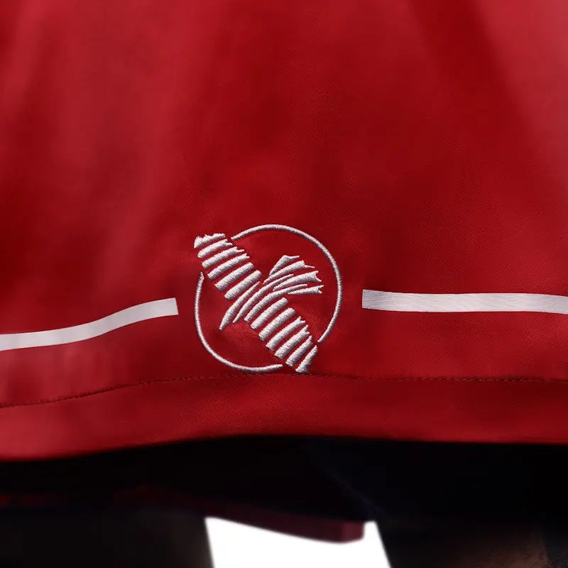 Close-up of the embroidered Hayabusa logo on red boxing shorts, positioned near the hem with white accent lines. The image highlights the fine stitching, smooth satin fabric, and clean finish, emphasizing durability, premium craftsmanship, and a lightweight feel for boxing training and gym use.