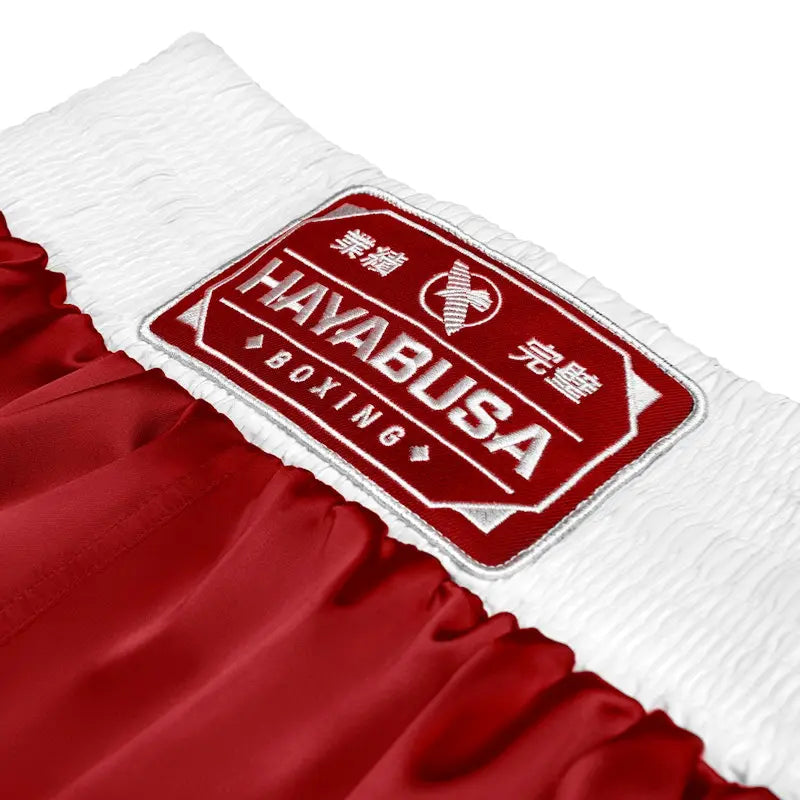 Close-up of the waistband patch on red Hayabusa boxing shorts, featuring the stitched Hayabusa Boxing logo on a wide white elastic waistband. The image highlights the premium embroidery, textured elastic fabric, and smooth red satin material, emphasizing durability, comfort, and a secure fit for boxing training and gym use.