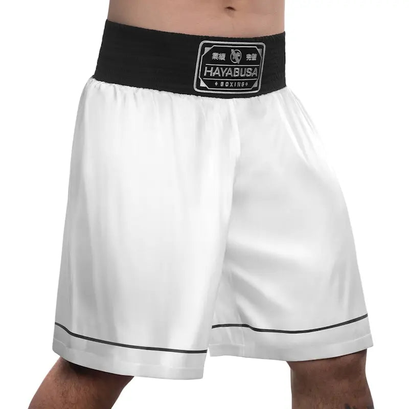 Front view of white Hayabusa boxing shorts featuring a wide black elastic waistband with the Hayabusa Boxing patch. The image shows the lightweight fabric, loose fit around the thighs, and clean black piping along the hem, designed for comfort, mobility, and stability during boxing training and gym workouts.