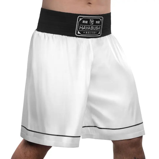 Front view of white Hayabusa boxing shorts featuring a wide black elastic waistband with the Hayabusa Boxing patch. The image shows the lightweight fabric, loose fit around the thighs, and clean black piping along the hem, designed for comfort, mobility, and stability during boxing training and gym workouts.