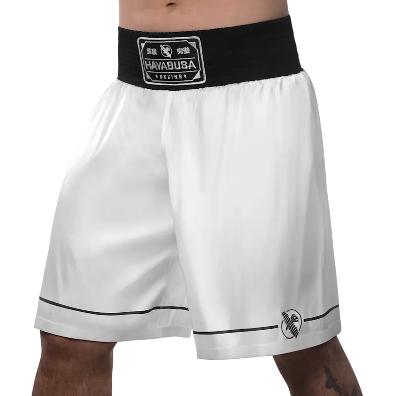 Front-left angle of white Hayabusa boxing shorts with a wide black elastic waistband and Hayabusa Boxing logo patch. The image highlights the loose, lightweight fit, smooth satin-like fabric, subtle black piping along the hem, and small Hayabusa logo on the leg, designed for freedom of movement and comfort during boxing training and gym sessions.