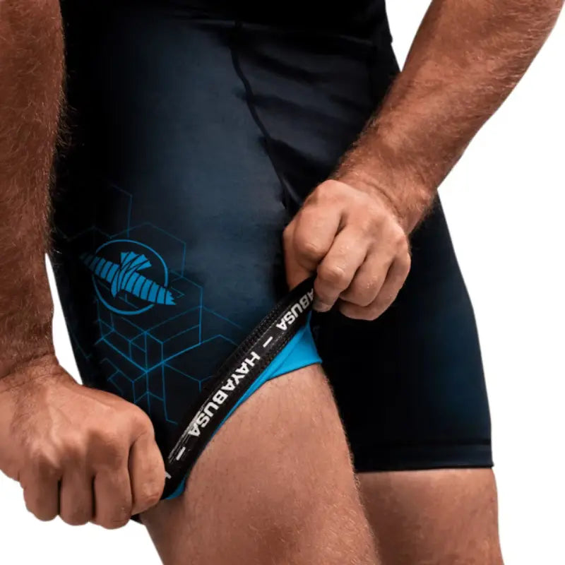 Close-up van zwarte Hayabusa compressieshort met blauwe details, waarbij de sporter de zoom optrekt met zichtbaar Hayabusa-logo.