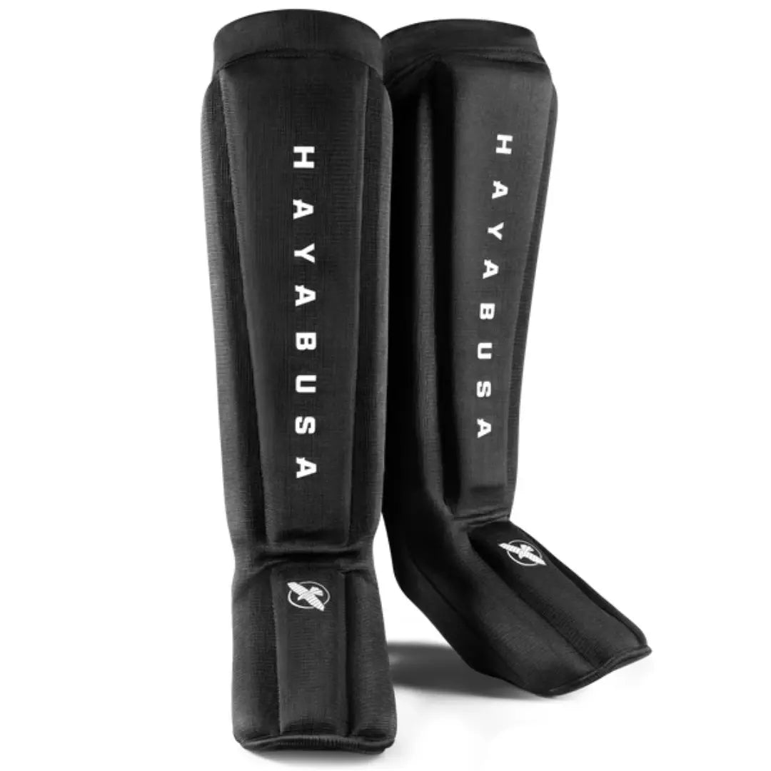 Productfoto van zwarte scheenbeschermers met voetbescherming en witte Hayabusa-logo’s. De Hayabusa Core Sleeved Scheenbeschermers Zwart zijn geschikt voor kickboksers, Muay Thai-sporters en MMA-beoefenaars die lichte bescherming en een strakke pasvorm zoeken tijdens training.