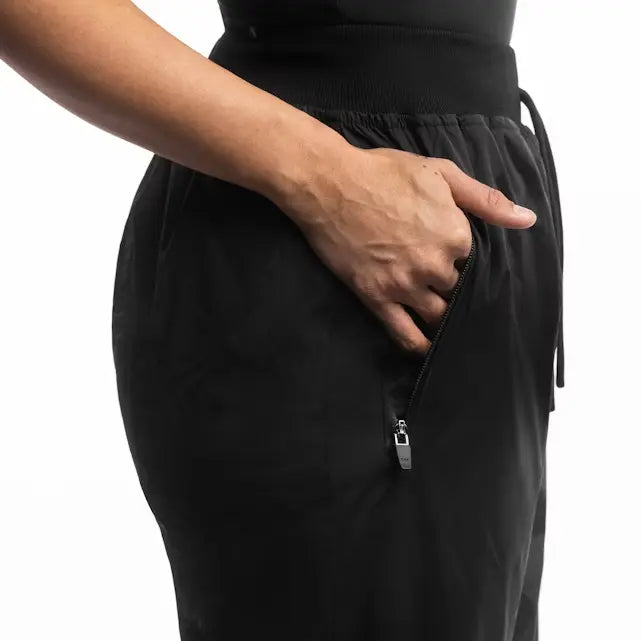 Een close-up toont de hand van een persoon in de zijzak van een zwarte saunapak-broek. De zak is voorzien van een metalen rits en een trekkoord bij de tailleband.