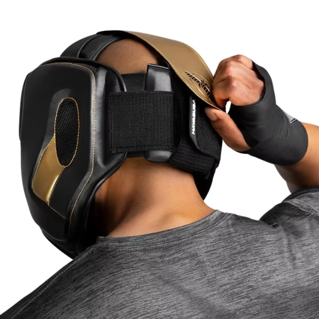 Sporter zet zwarte en gouden Hayabusa hoofdbeschermer vast met klittenband tijdens bokstraining.