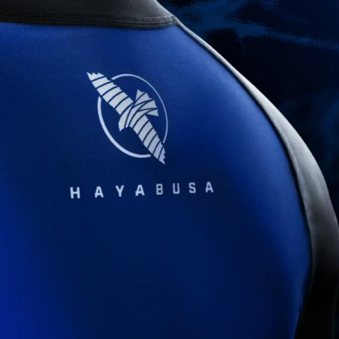 Close-up van het Hayabusa-logo op de bovenrug van de blauwe rashguard. De witte opdruk steekt scherp af tegen de diepblauwe stof. De foto laat de printkwaliteit en het gladde, rekbare materiaal duidelijk naar voren komen.