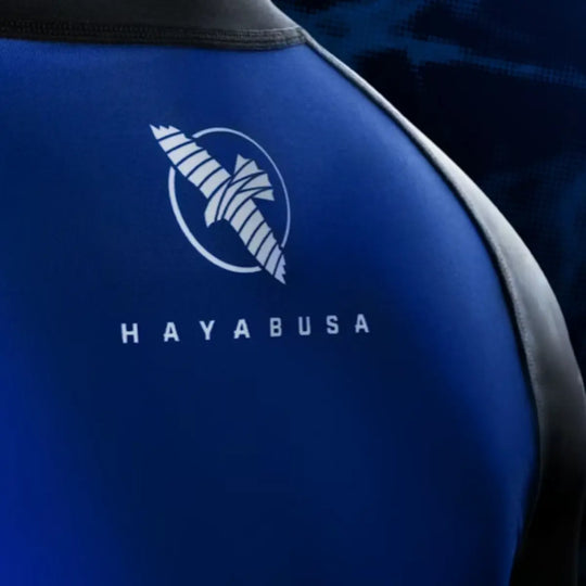 Close-up van het Hayabusa-logo op de bovenrug van de blauwe rashguard. De witte opdruk steekt scherp af tegen de diepblauwe stof. De foto laat de printkwaliteit en het gladde, rekbare materiaal duidelijk naar voren komen.