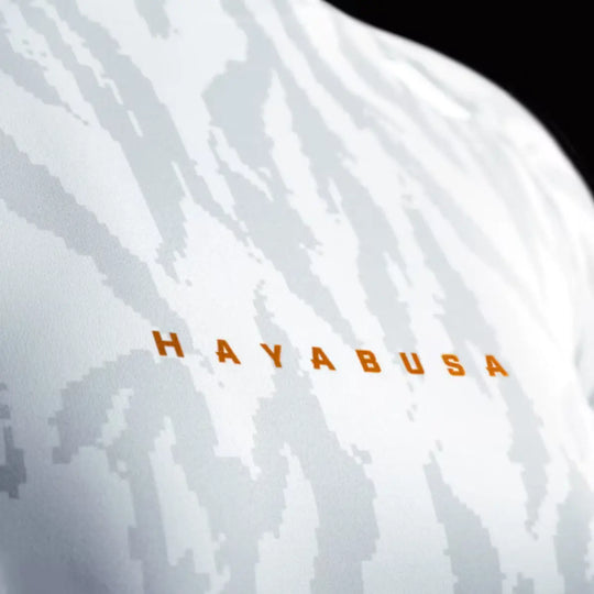 Close-up van witte Hayabusa rashguard met grijze camouflageprint en oranje merklogo op de borst.