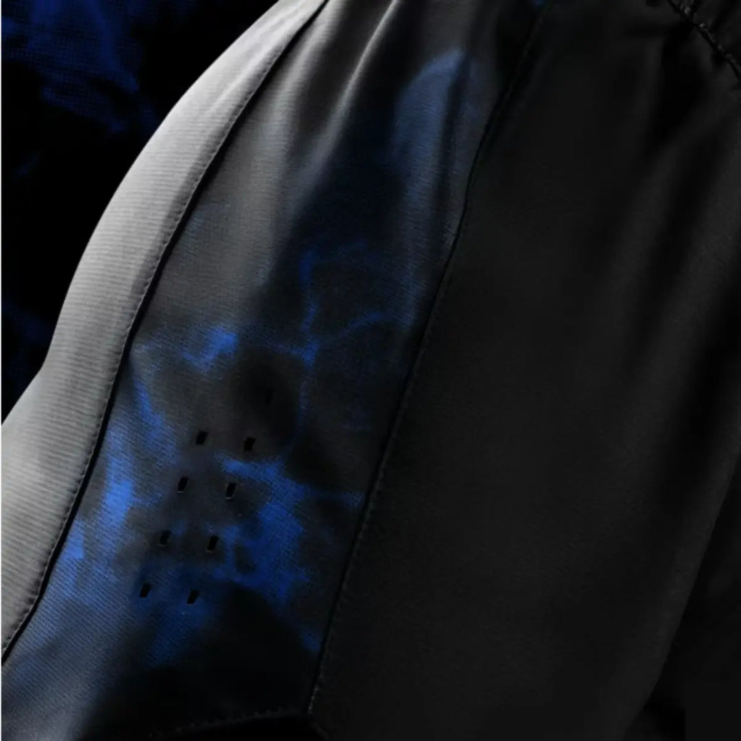Zijaanzicht van het blauwe camouflagepaneel met ventilatiegaatjes op de shorts. De textuur van het materiaal en de perforaties laten luchtcirculatie zien voor extra comfort. Het zwarte hoofddeel van de short contrasteert sterk met de blauwe accenten. De foto benadrukt functionaliteit en premium afwerking.