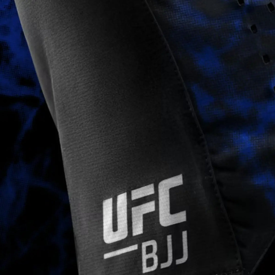 Close-up van het UFC BJJ-logo op het been van de shorts. Het witte logo steekt scherp af tegen het zwarte materiaal. Het blauwe camouflagepaneel naast het logo geeft de shorts een sportieve uitstraling. De foto benadrukt de afwerking en printkwaliteit.
