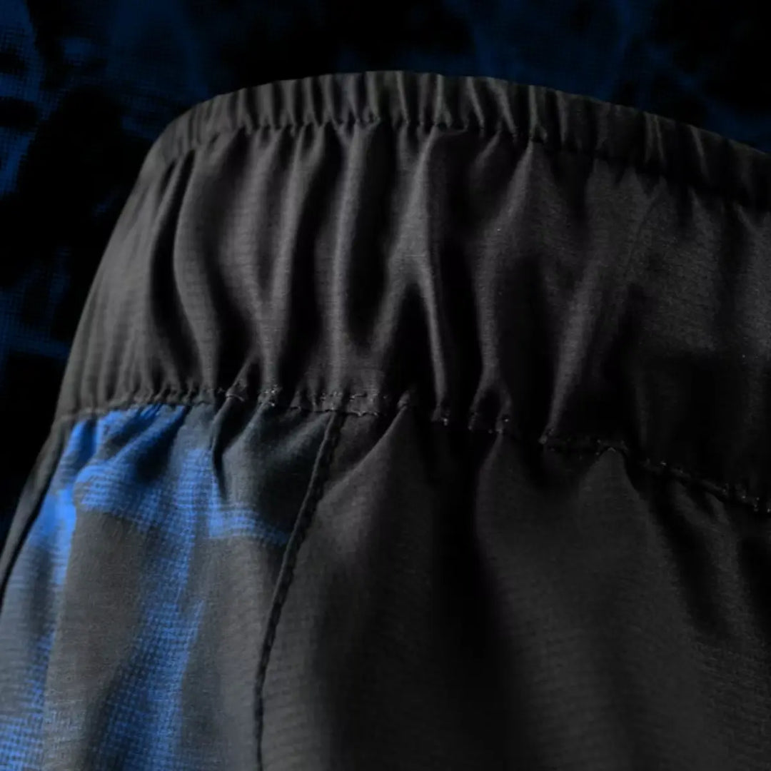 Close-up van de elastische tailleband van de UFC BJJ Women’s Shorts in zwart. De plooien en stiknaden laten de stevige constructie en rekbaarheid zien. Het blauwe zijpaneel komt subtiel in beeld. De donkere achtergrond benadrukt het materiaalcontrast.