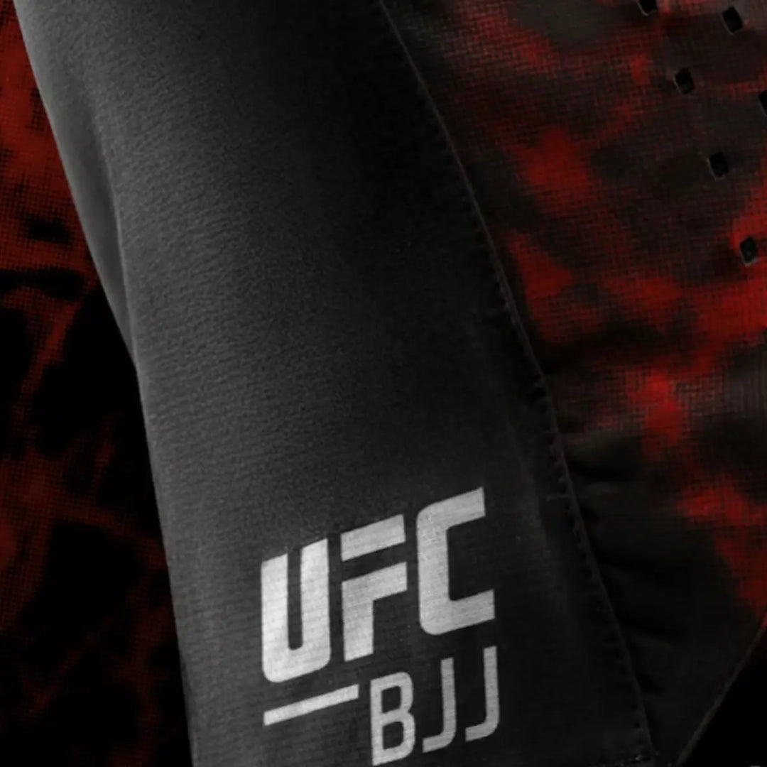 Close-up van het witte UFC BJJ-logo op het been van de shorts. Het logo steekt helder af tegen het zwarte textiel. Het rode camouflagepaneel zorgt voor een krachtig visueel effect. De foto focust op de printkwaliteit en afwerking.