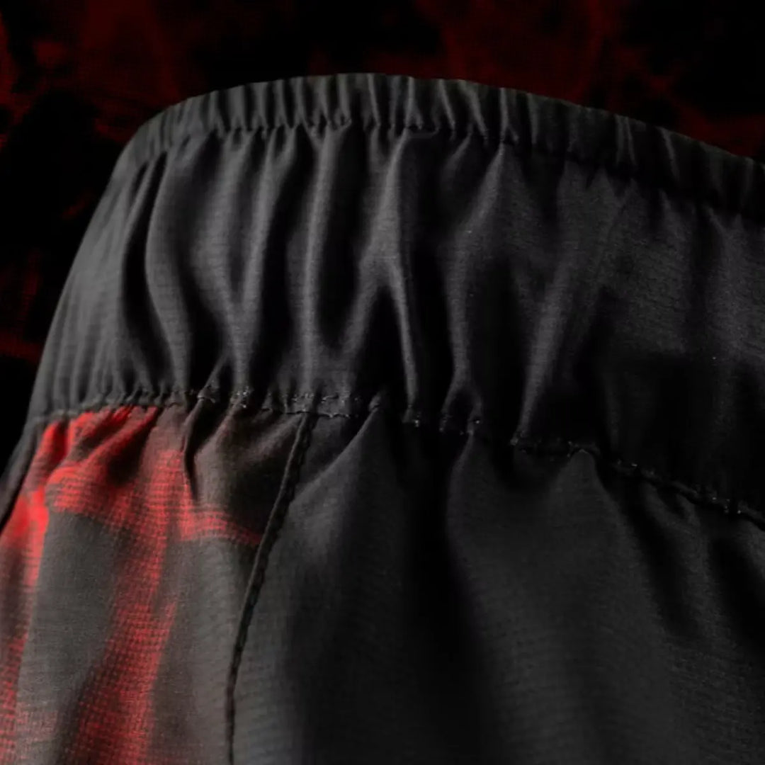 Detail van de elastische tailleband van de UFC BJJ Women’s Shorts. De plooien en naden tonen de rekbaarheid en stevigheid van het materiaal. Het rode zijpaneel komt subtiel in beeld. De achtergrond met rode accenten benadrukt de sportieve look.