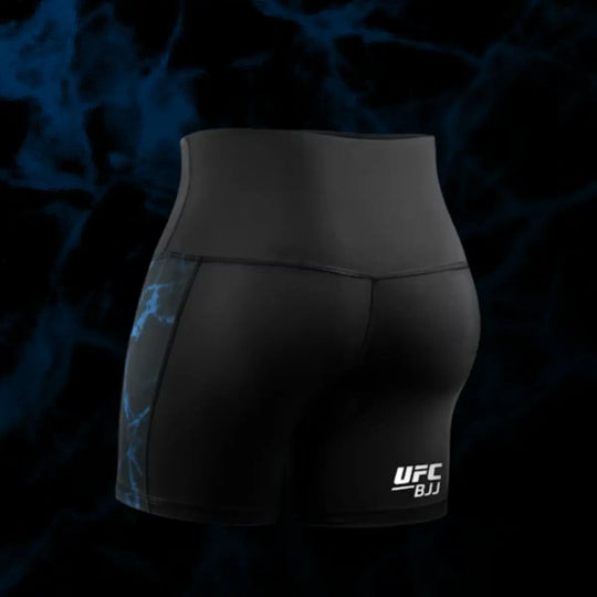 Achteraanzicht van de zwarte UFC BJJ Women’s Vale Tudo Shorts, met blauwe zijpanelen en witte logo-opdruk. De strakke compressiepasvorm benadrukt ondersteuning en bewegingsvrijheid. De naden zijn netjes afgewerkt en de shorts sluiten anatomisch aan. De donkere achtergrond versterkt de premium uitstraling.